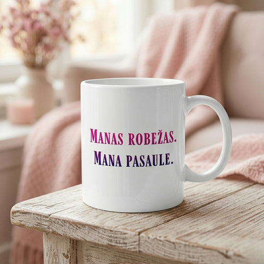 Balta 330ml keramikas krūze ar tekstu "MANAS ROBEŽAS. MANA PASAULE." purpursarkanā un tumši zilā krāsā. Krūze novietota uz gaiša koka galda, fonā mājīgs interjers ar rozā pledu un ziediem maigos toņos.