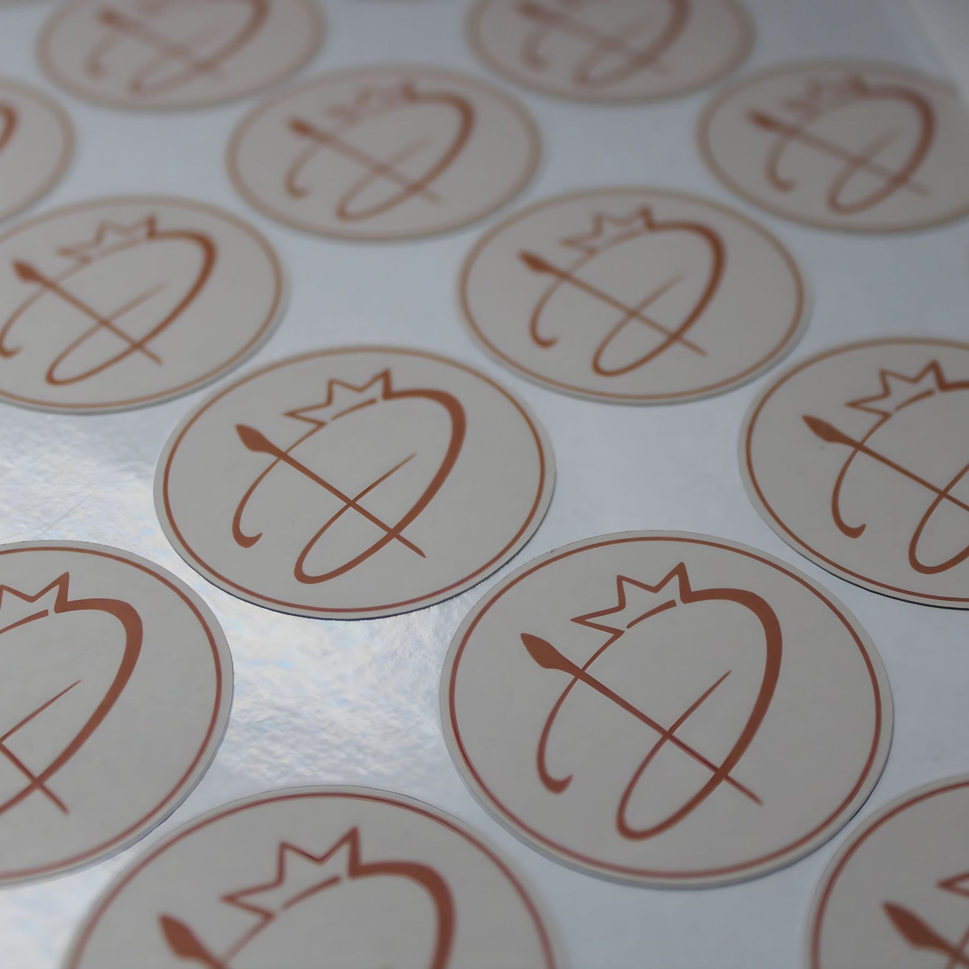 Apaļu, glancētu uzlīmju loksne tuvplānā. Uzlīmēm ir maigi rozā fons ar bronzas krāsas logotipu – stilizētu burtu, ko papildina kroņa un otas motīvs. Virsma atstaro gaismu, norādot uz "glossy" materiāla tekstūru.