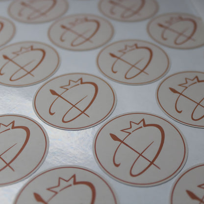 Apaļu, glancētu uzlīmju loksne tuvplānā. Uzlīmēm ir maigi rozā fons ar bronzas krāsas logotipu – stilizētu burtu, ko papildina kroņa un otas motīvs. Virsma atstaro gaismu, norādot uz "glossy" materiāla tekstūru.