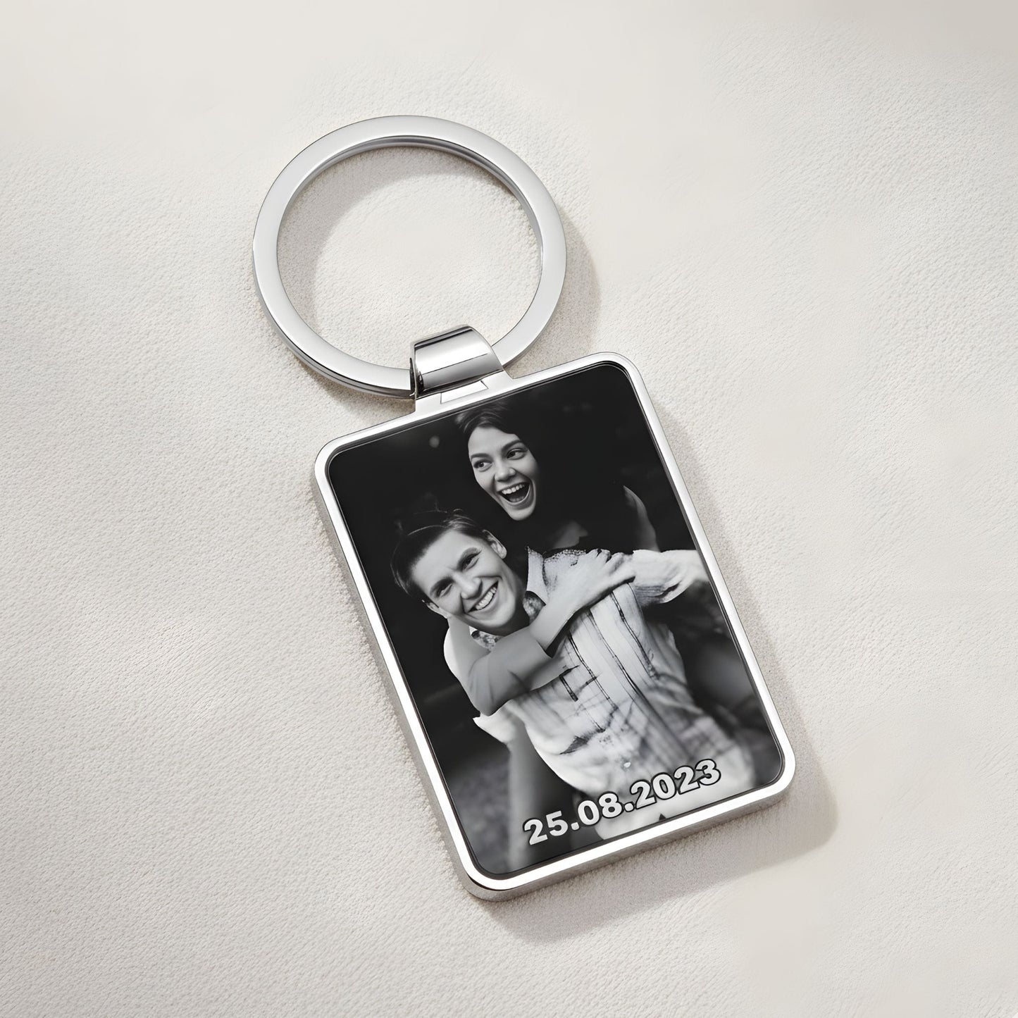 Personalizēts taisnstūra formas metāla atslēgu piekariņš ar melnbaltu fotogrāfiju un datumu uz neitrāla, teksturēta smilšu krāsas fona. Skats no augšas.