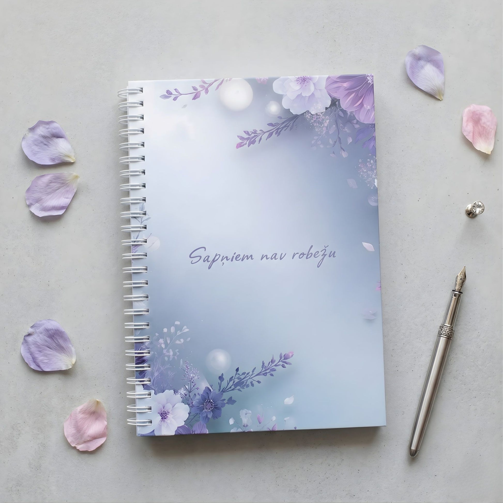 Minimālistiska dizaina klade ar ziedu motīviem maigi zilos un violetos toņos, uzraksts "Sapņiem nav robežu", novietota uz gaiši pelēka fona blakus elegantai pildspalvai un ziedlapiņām.
