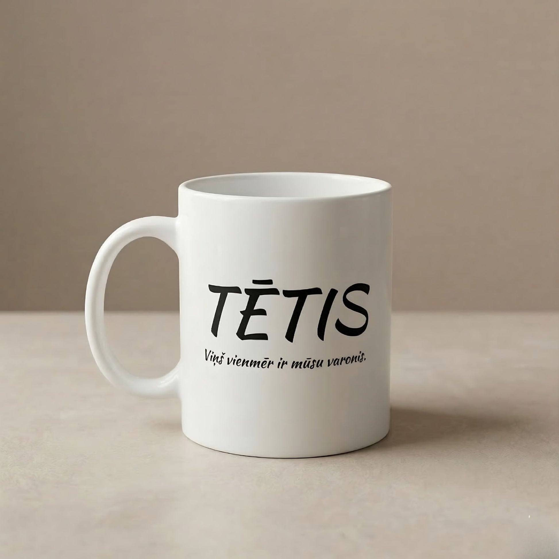 Balta, tukša keramikas krūze novietota uz minimālistiska, silta bēša fona. Uz krūzes ir melns teksts latviešu valodā: liels uzraksts "TĒTIS" un apakšā mazāks teksts kursīvā "Viņš vienmēr ir mūsu varonis.".