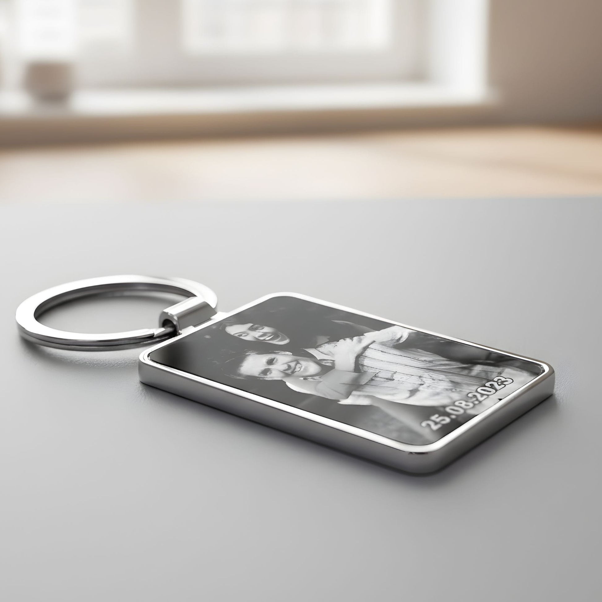 Personalizēts metāla atslēgu piekariņš ar melnbaltu fotoattēlu, novietots uz pelēka paliktņa uz koka galda.