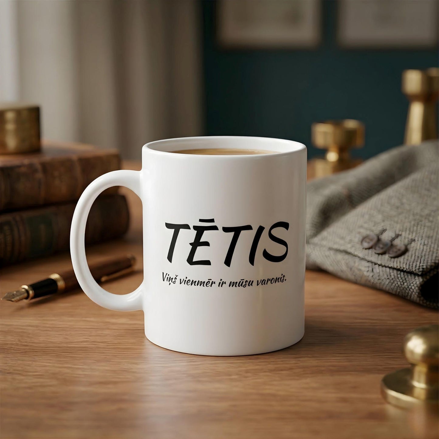 Balta keramikas krūze ar melnu tekstu "TĒTIS. Viņš vienmēr ir mūsu varonis." uz mājīga darba galda fona ar ādas grāmatu un spalvaskātu.