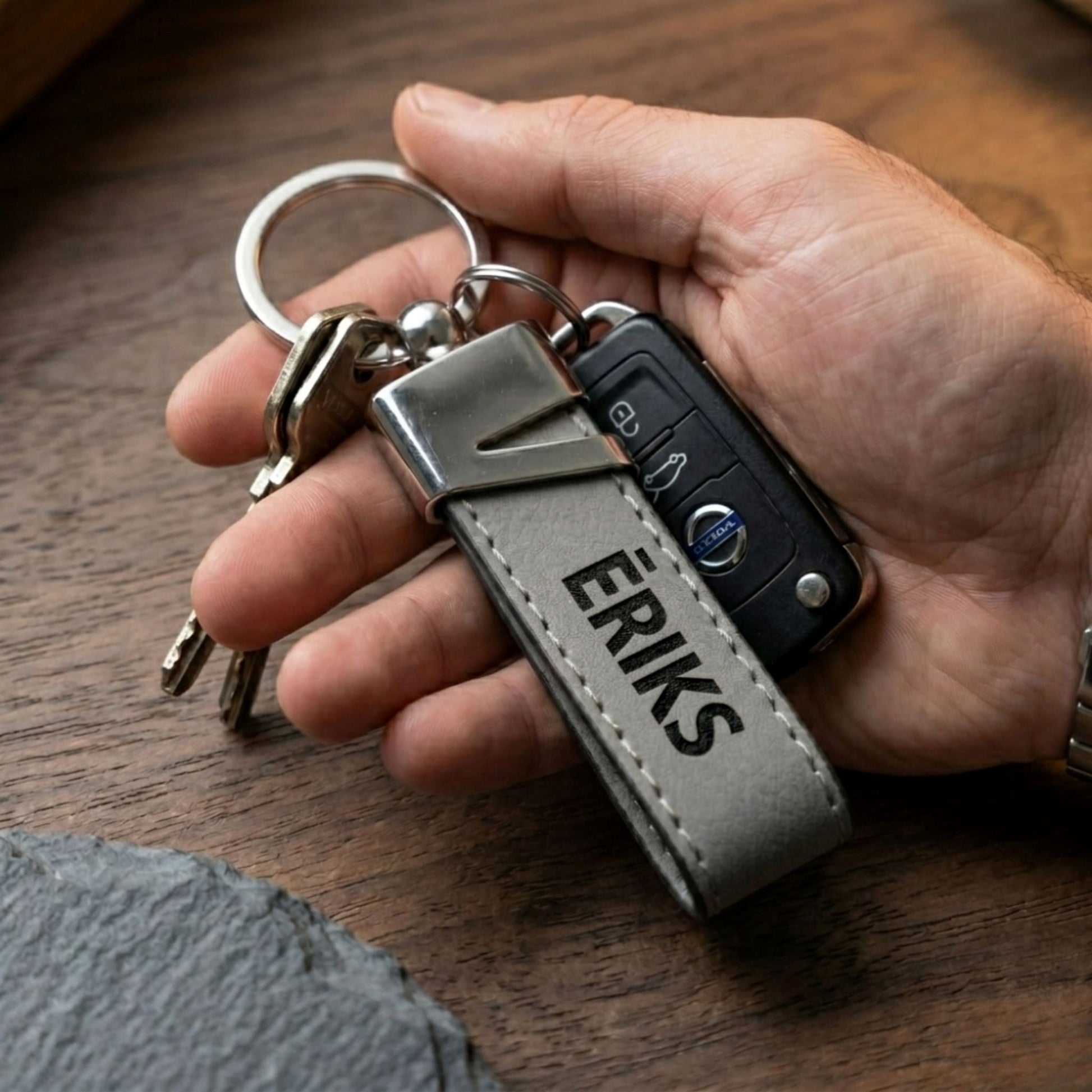 Personalizēts pelēks ādas atslēgu piekariņš ar vārdu "ĒRIKS" un automašīnas atslēgām cilvēka rokā, demonstrējot tā izmēru un lietošanu.