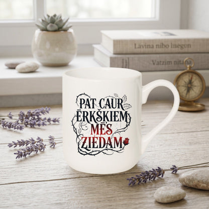 Balta noapaļotas formas krūze ar latviešu valodas motivējošu uzrakstu "Pat caur ērkšķiem mēs ziedam" un rožu grafiku, fona dekorācijas ar lavandu un grāmatām.