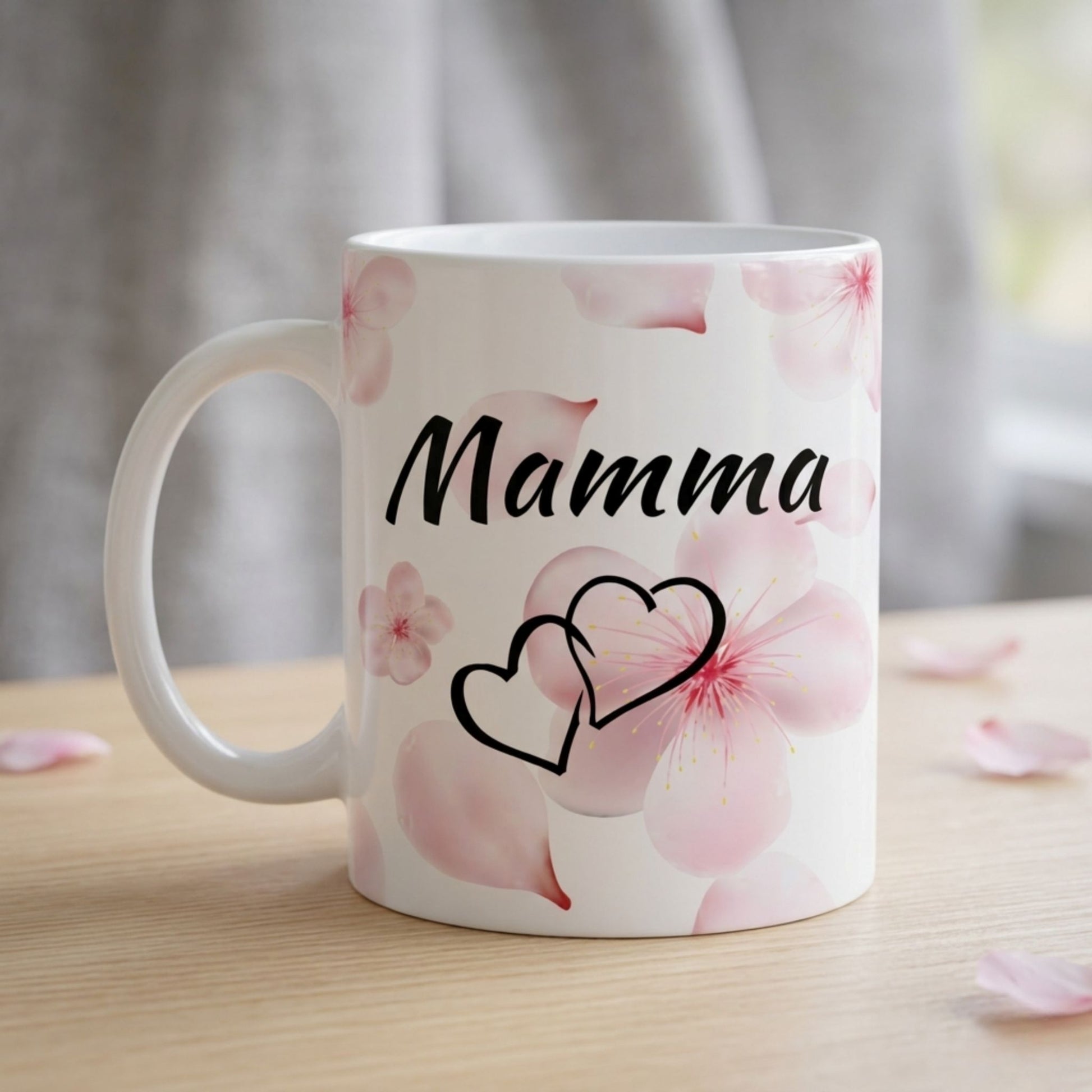 Balta 330 ml keramikas krūze ar uzrakstu "Mamma", divu siržu simbolu un maigi rozā sakuras ziedlapu dizainu uz koka galda fona.