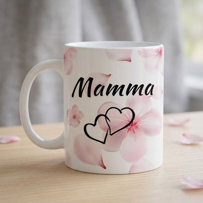 Balta 330 ml keramikas krūze ar uzrakstu "Mamma", divu siržu simbolu un maigi rozā sakuras ziedlapu dizainu uz koka galda fona.