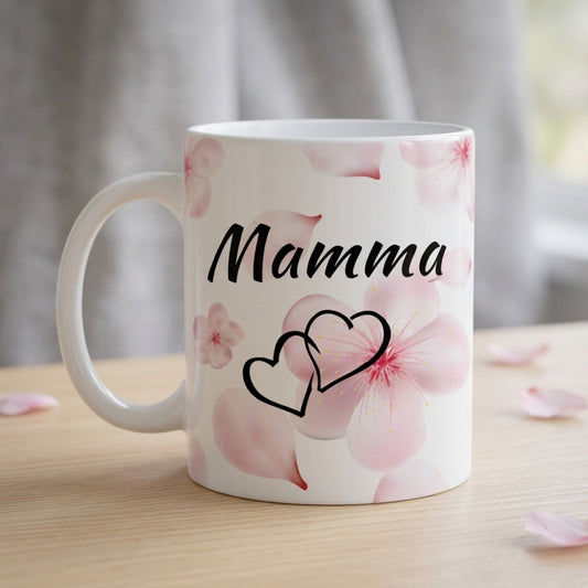 Balta 330 ml keramikas krūze ar uzrakstu "Mamma", divu siržu simbolu un maigi rozā sakuras ziedlapu dizainu uz koka galda fona.