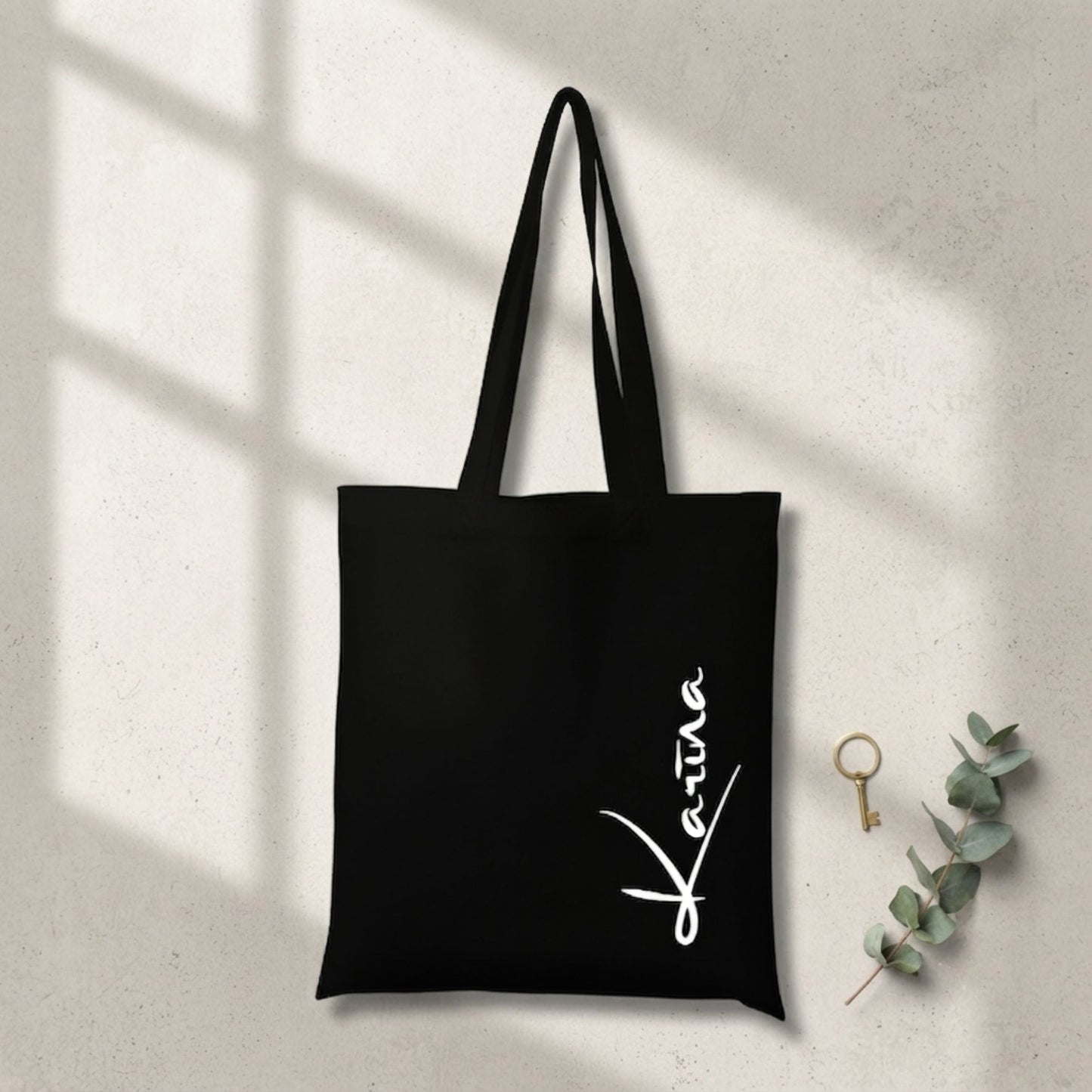 Melna, izturīga auduma canvas soma ar baltu, vertikālu kaligrāfisku uzrakstu "Karina". Soma novietota uz gaišas, teksturētas virsmas, ko šķērso reālistiska loga rāmja ēna, blakus redzams zelta gredzens un eikalipta zars.