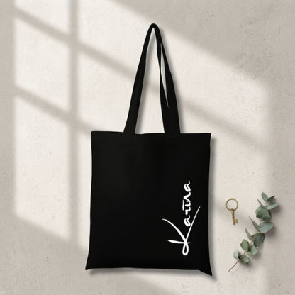 Melna, izturīga auduma canvas soma ar baltu, vertikālu kaligrāfisku uzrakstu "Karina". Soma novietota uz gaišas, teksturētas virsmas, ko šķērso reālistiska loga rāmja ēna, blakus redzams zelta gredzens un eikalipta zars.