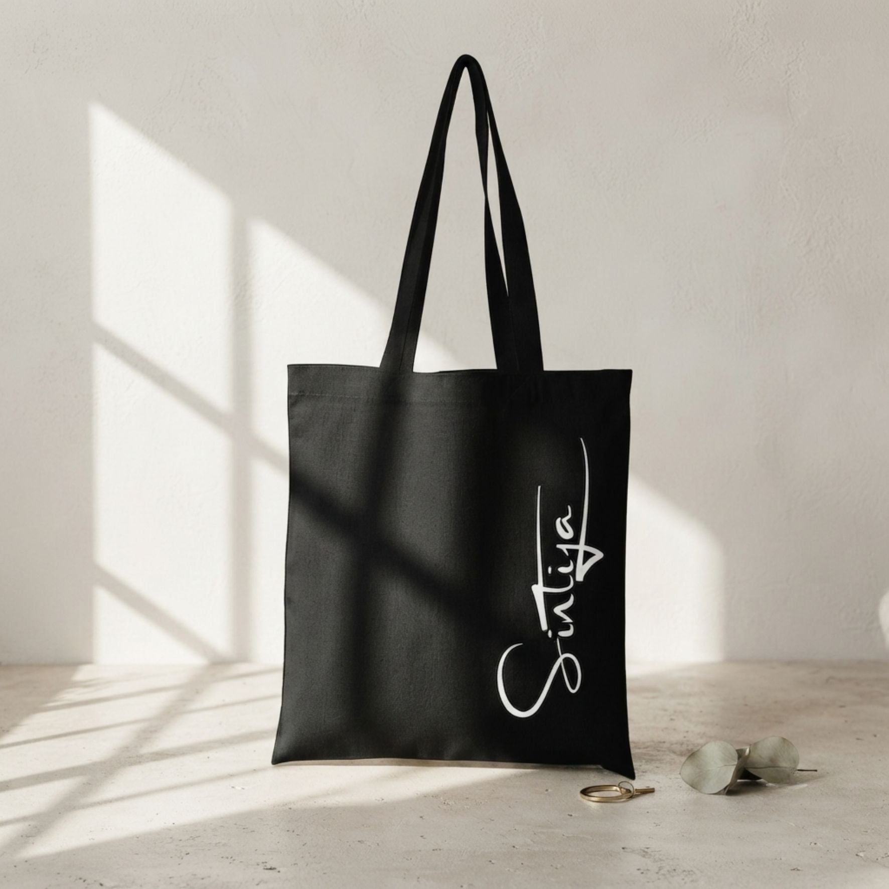 Melna, izturīga auduma canvas soma ar baltu, vertikālu kaligrāfisku uzrakstu "Sintija". Soma novietota uz gaišas, teksturētas virsmas, ko šķērso reālistiska loga rāmja ēna, blakus redzams zelta gredzens un eikalipta zars.