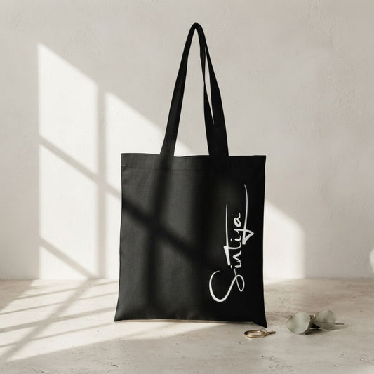 Melna, izturīga auduma canvas soma ar baltu, vertikālu kaligrāfisku uzrakstu "Sintija". Soma novietota uz gaišas, teksturētas virsmas, ko šķērso reālistiska loga rāmja ēna, blakus redzams zelta gredzens un eikalipta zars.