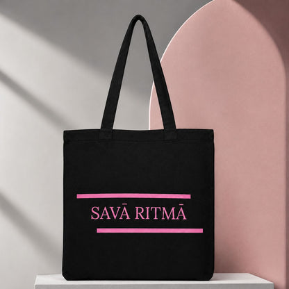 Melna kanvas soma “Savā ritmā” ar rozā apdruku uz pelēka un rozā fona.