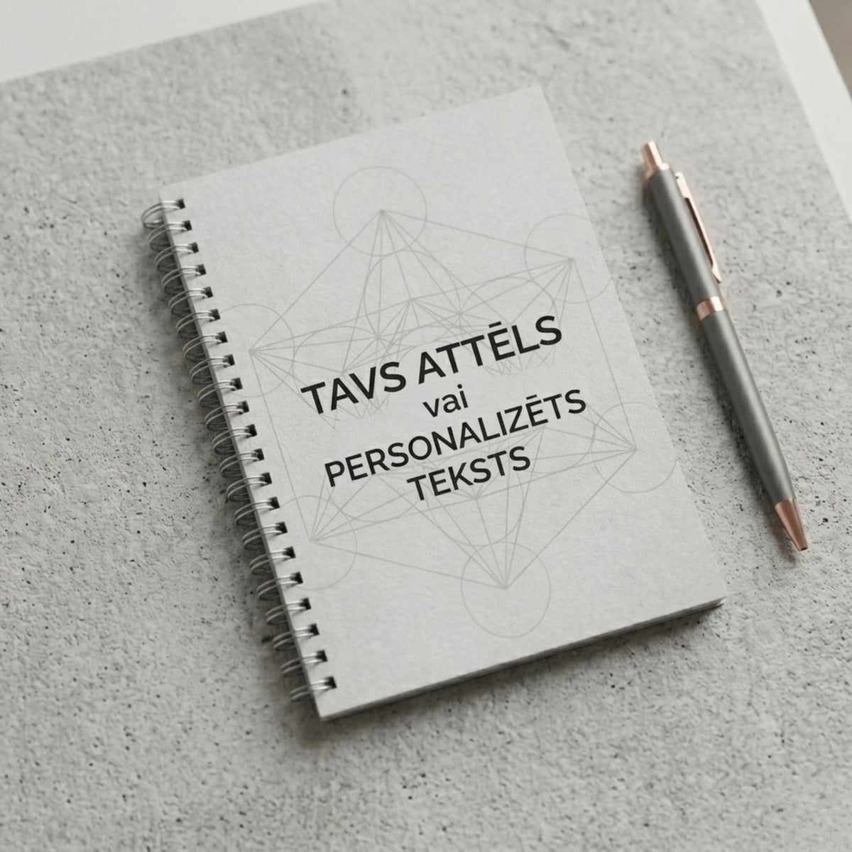 Minimālistiska pelēka piezīmju klade ar spirāli un uzrakstu "Tavs attēls vai personalizēts teksts" uz betona tekstūras fona ar pildspalvu.