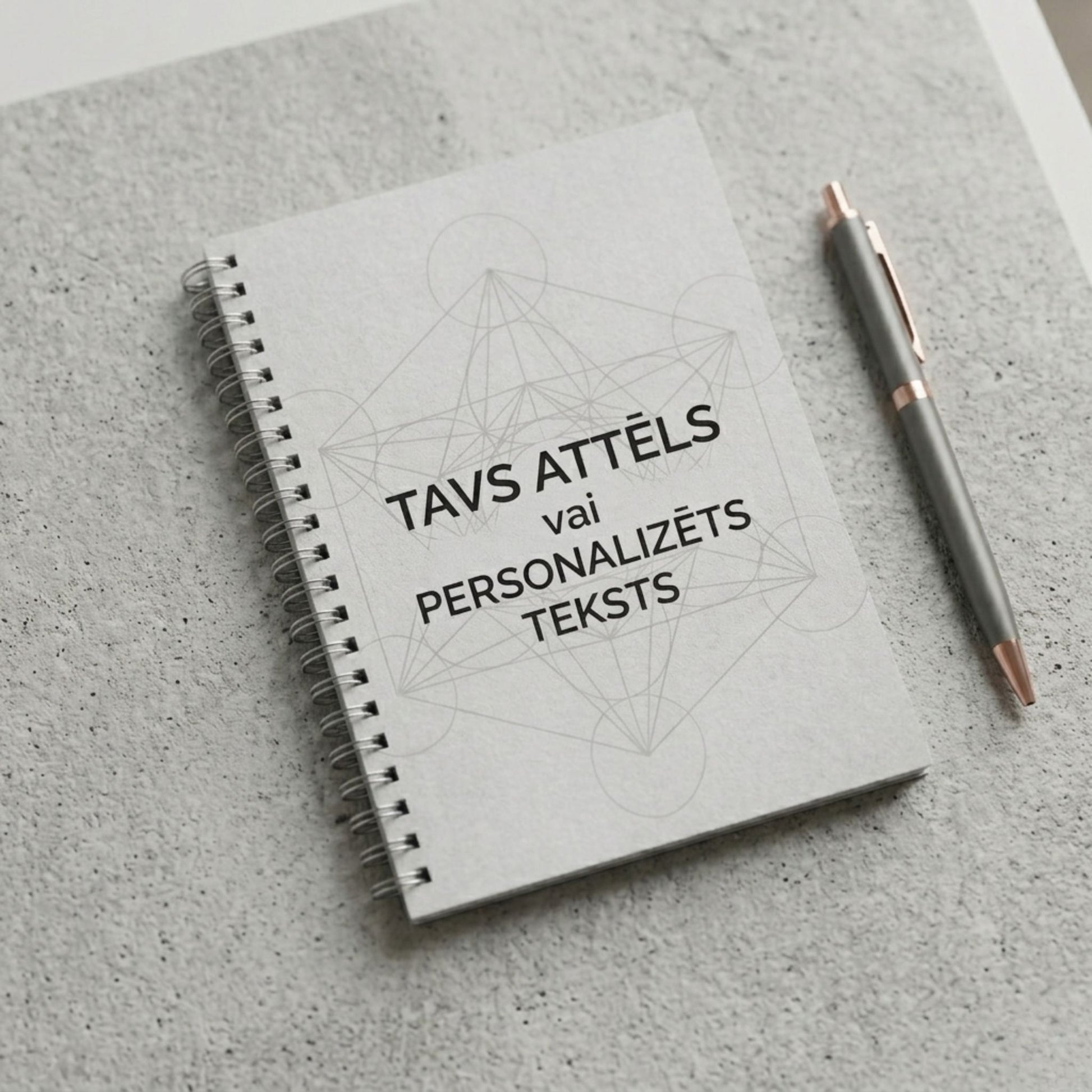 Minimālistiska pelēka piezīmju klade ar spirāli un uzrakstu "Tavs attēls vai personalizēts teksts" uz betona tekstūras fona ar pildspalvu.