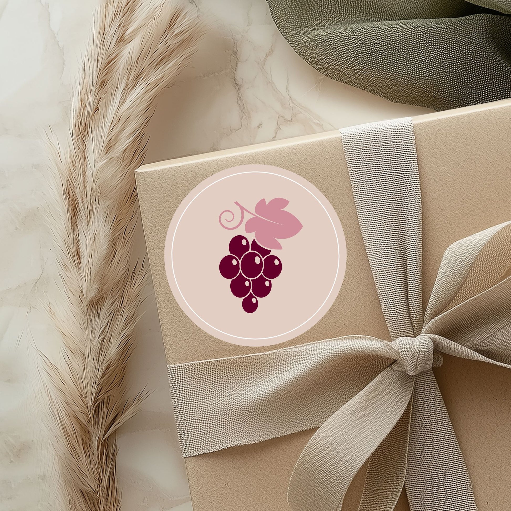 Tuvplāna attēls ar gaiši brūnu dāvanu kasti, kas pārsieta ar elegantu lenti. Uz kastes uzlīmēta apaļa, maigi rozā uzlīme ar vīnogu ķekara logo. Blakus redzama pūkaina dekoratīvā smilga uz marmora virsmas fona.