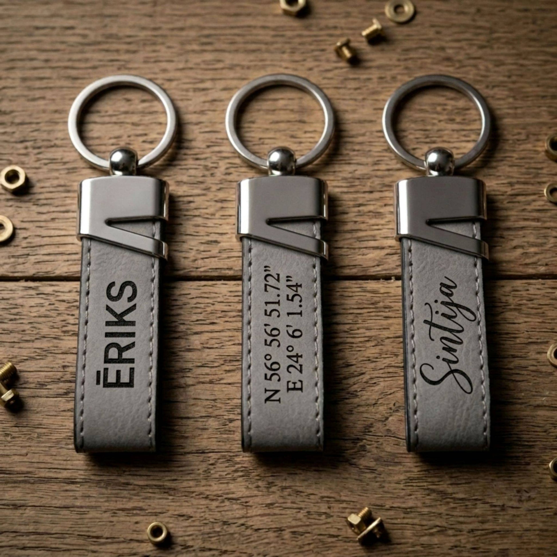 Trīs dažādi personalizēti pelēkas ādas atslēgu piekariņi ar melnu gravējumu: vārds "ĒRIKS", ģeogrāfiskās koordinātas un vārds "Sintija" kaligrāfiskā fontā.
