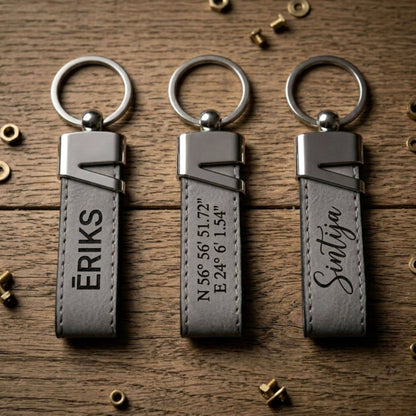 Trīs dažādi personalizēti pelēkas ādas atslēgu piekariņi ar melnu gravējumu: vārds "ĒRIKS", ģeogrāfiskās koordinātas un vārds "Sintija" kaligrāfiskā fontā.