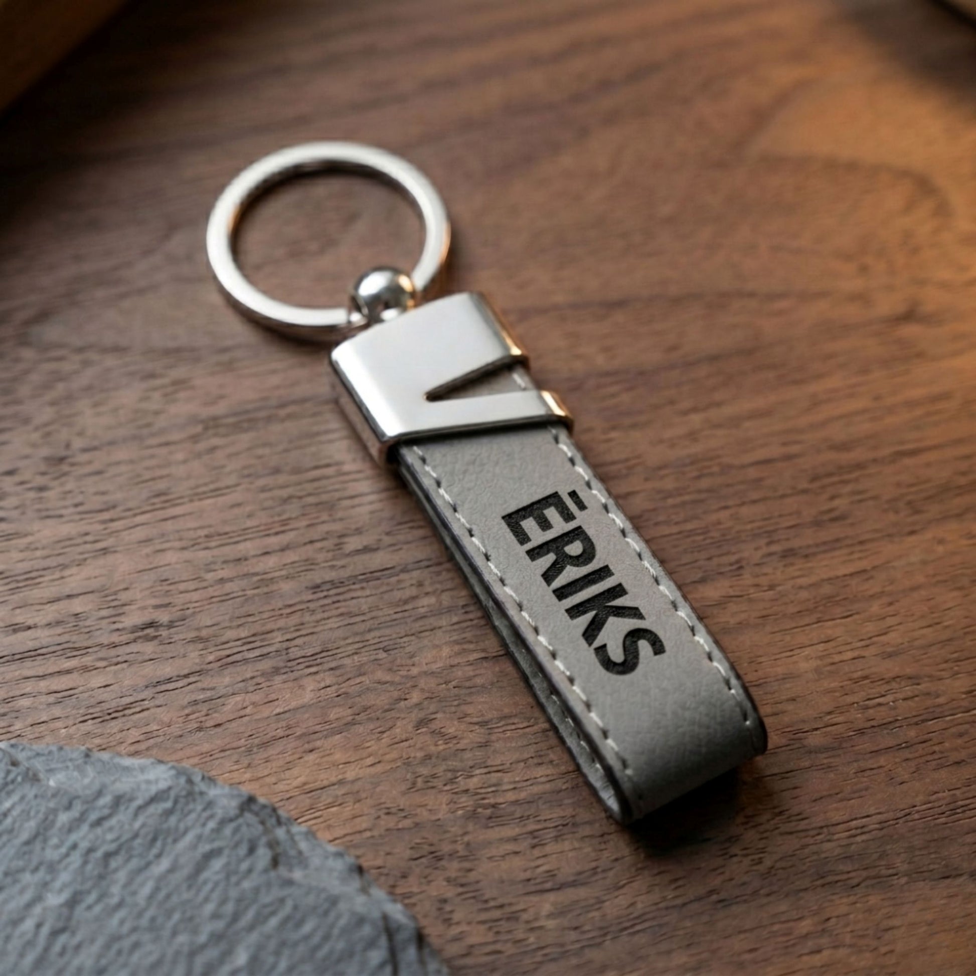 Minimālistisks pelēkas ādas atslēgu piekariņš ar melnu lāzera gravējumu "ĒRIKS" uz tumša koka fona.