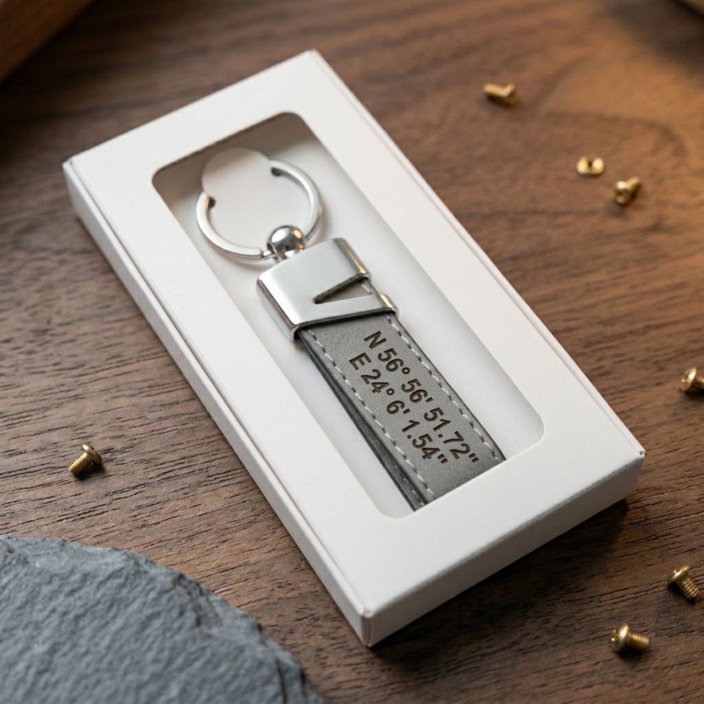 Personalizēts pelēks ādas atslēgu piekariņš ar iegravētām koordinātām, ievietots elegantā baltā dāvanu kastītē ar lodziņu.