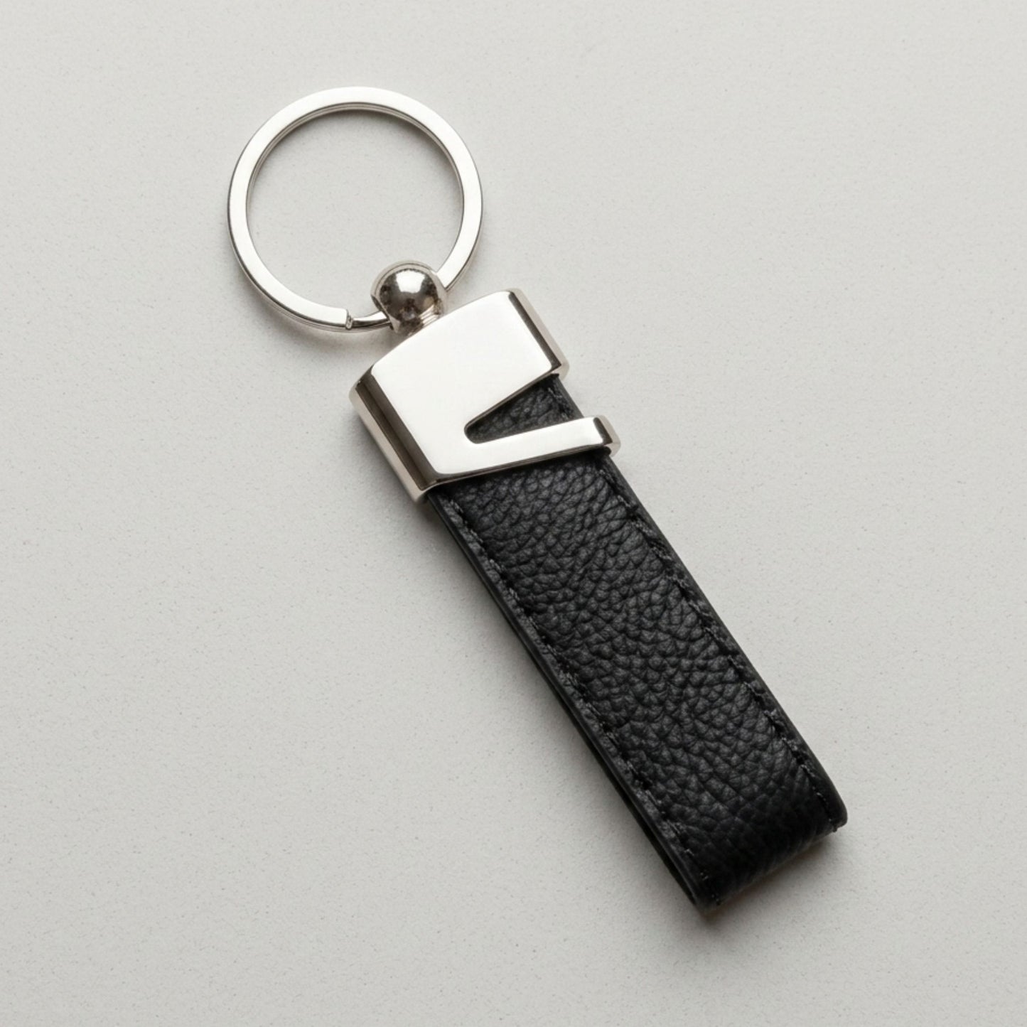 Melnas ādas un metāla atslēgu piekariņa tuvplāns ar precīzu un skaidru personalizētu gravējumu "MAREKS" uz pelēka fona.
